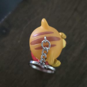 Op de afbeelding: Een kleine oranje figuur-sleutelhanger met bruine strepen op het hoofd en een zilveren metalen ketting met een ronde ring aan de bovenkant. Het figuurtje heeft een puntig oor en een glad, afgerond oppervlak, gezien vanuit vogelperspectief.