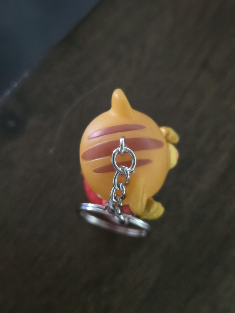 Op de afbeelding: Een kleine oranje figuur-sleutelhanger met bruine strepen op het hoofd en een zilveren metalen ketting met een ronde ring aan de bovenkant. Het figuurtje heeft een puntig oor en een glad, afgerond oppervlak, gezien vanuit vogelperspectief.