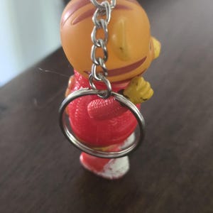 Op de afbeelding: Een kleine oranje tijgerfiguur sleutelhanger met bruine strepen, gekleed in een rode trui met textuur en witte schoenen. Het figuurtje heeft een zilverkleurige metalen ketting en een ronde sleutelring bevestigd aan de bovenkant van het hoofd.