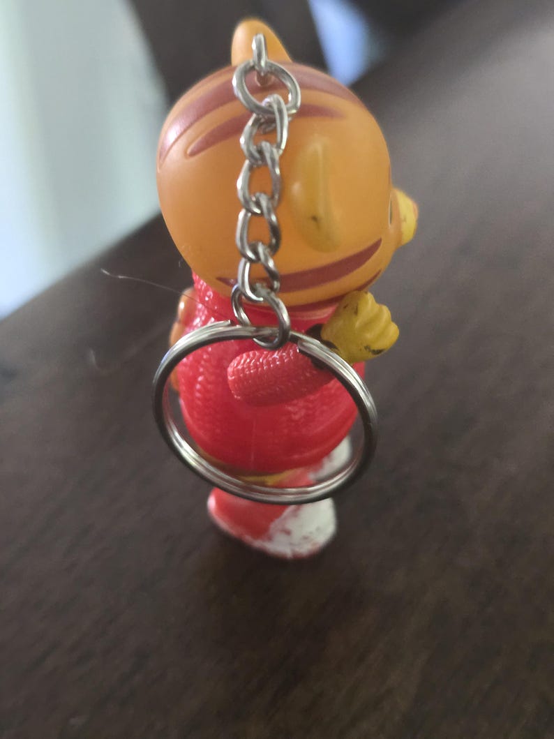 Op de afbeelding: Een kleine oranje tijgerfiguur sleutelhanger met bruine strepen, gekleed in een rode trui met textuur en witte schoenen. Het figuurtje heeft een zilverkleurige metalen ketting en een ronde sleutelring bevestigd aan de bovenkant van het hoofd.