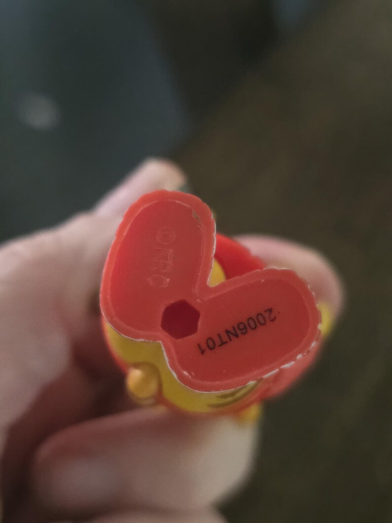 Op de afbeelding: De onderkant van een felrode plastic voet van een figuurtje met een zeshoekig gat in het midden. De tekst "&copy;FRC" staat in reli&euml;f aan de linkerkant en de alfanumerieke code "2006NT01" is met zwarte inkt aan de rechterkant van de zool gedrukt.