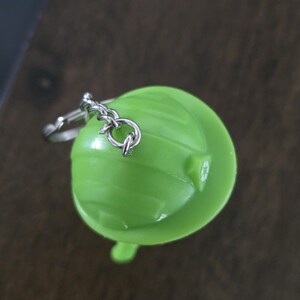May include: A bright lime green plastic hard hat keychain featuring a small silver metal chain and ring attachment. The top of the hard hat displays a subtle, molded recycling symbol on the side, with a smooth, rounded surface and horizontal ridges.