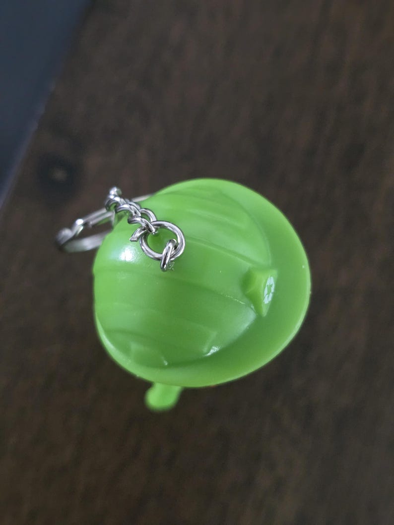 May include: A bright lime green plastic hard hat keychain featuring a small silver metal chain and ring attachment. The top of the hard hat displays a subtle, molded recycling symbol on the side, with a smooth, rounded surface and horizontal ridges.