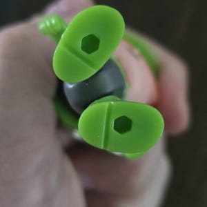 May include: A pair of bright lime green plastic feet from a small figurine. Each foot features a smooth, rounded sole with a distinct hexagonal hole centered in the middle, designed for mounting or display purposes on a compatible stand.