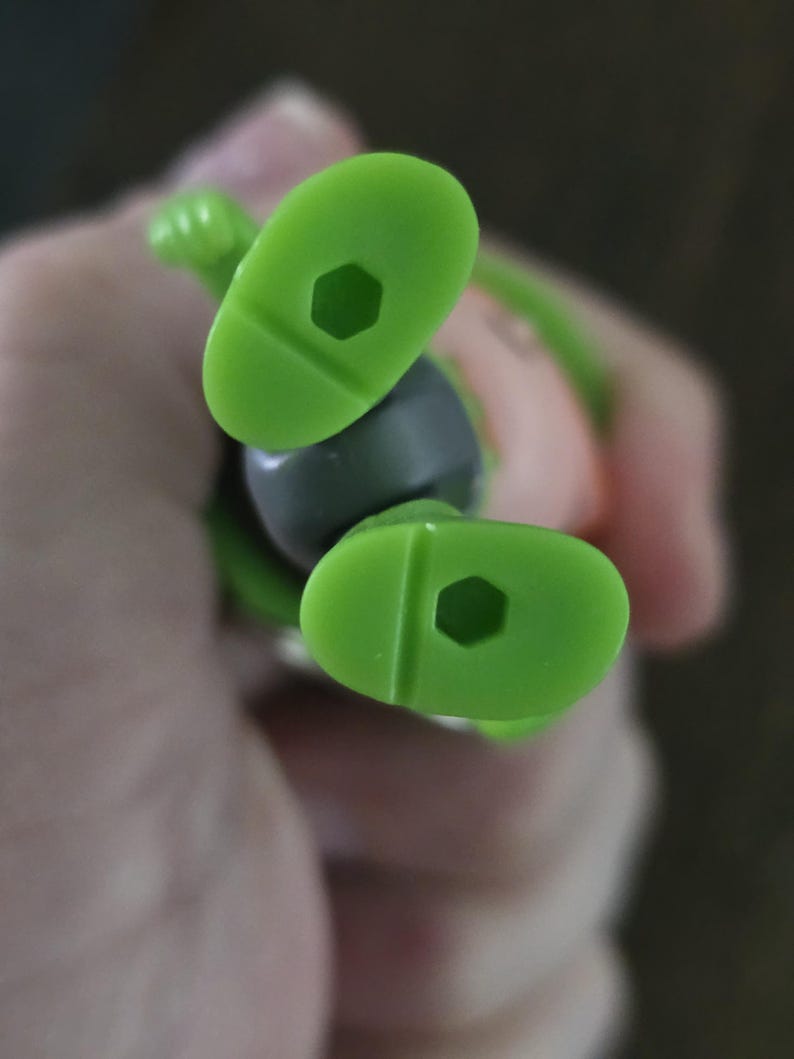 May include: A pair of bright lime green plastic feet from a small figurine. Each foot features a smooth, rounded sole with a distinct hexagonal hole centered in the middle, designed for mounting or display purposes on a compatible stand.