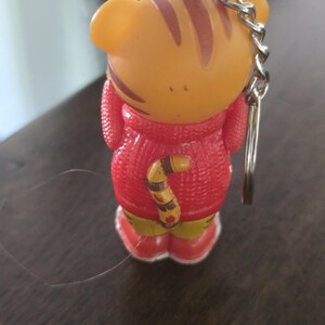 Op de afbeelding: Een kleine tijgerfiguur sleutelhanger met een oranje kop met bruine strepen, gekleed in een rode trui met textuur en rode schoenen. Het personage heeft een gestreepte staart aan de achterkant en een zilveren metalen ketting aan de bovenkant.