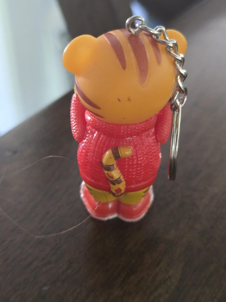 Op de afbeelding: Een kleine tijgerfiguur sleutelhanger met een oranje kop met bruine strepen, gekleed in een rode trui met textuur en rode schoenen. Het personage heeft een gestreepte staart aan de achterkant en een zilveren metalen ketting aan de bovenkant.