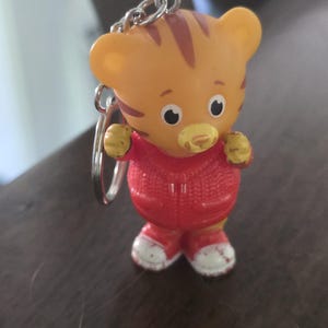 Op de afbeelding: Een kleine sleutelhanger in de vorm van een oranje tijgerfiguurtje met bruine strepen, een gele snuit en grote zwart-witte ogen. De tijger draagt een felrood vest met rits en een textuur van gebreid patroon, en witte sneakers met een rode rand.