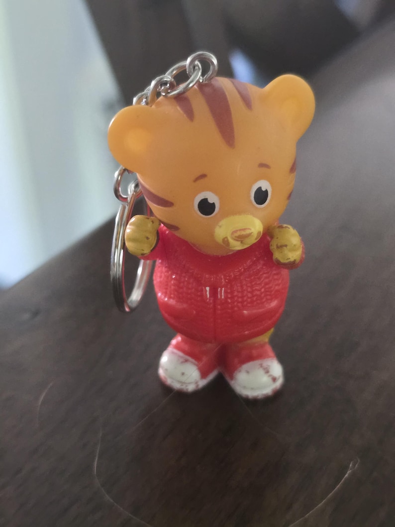 Op de afbeelding: Een kleine sleutelhanger in de vorm van een oranje tijgerfiguurtje met bruine strepen, een gele snuit en grote zwart-witte ogen. De tijger draagt een felrood vest met rits en een textuur van gebreid patroon, en witte sneakers met een rode rand.