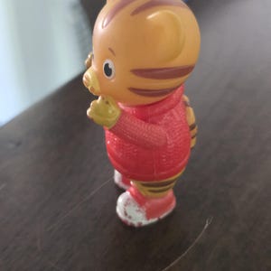 Op de afbeelding: Een klein oranje tijgerfiguurtje met bruine strepen, gekleed in een rode trui en rode schoenen met witte accenten. Het personage heeft een ronde kop, puntige oren en een metalen ring aan de bovenkant van het hoofd voor gebruik als sleutelhanger.