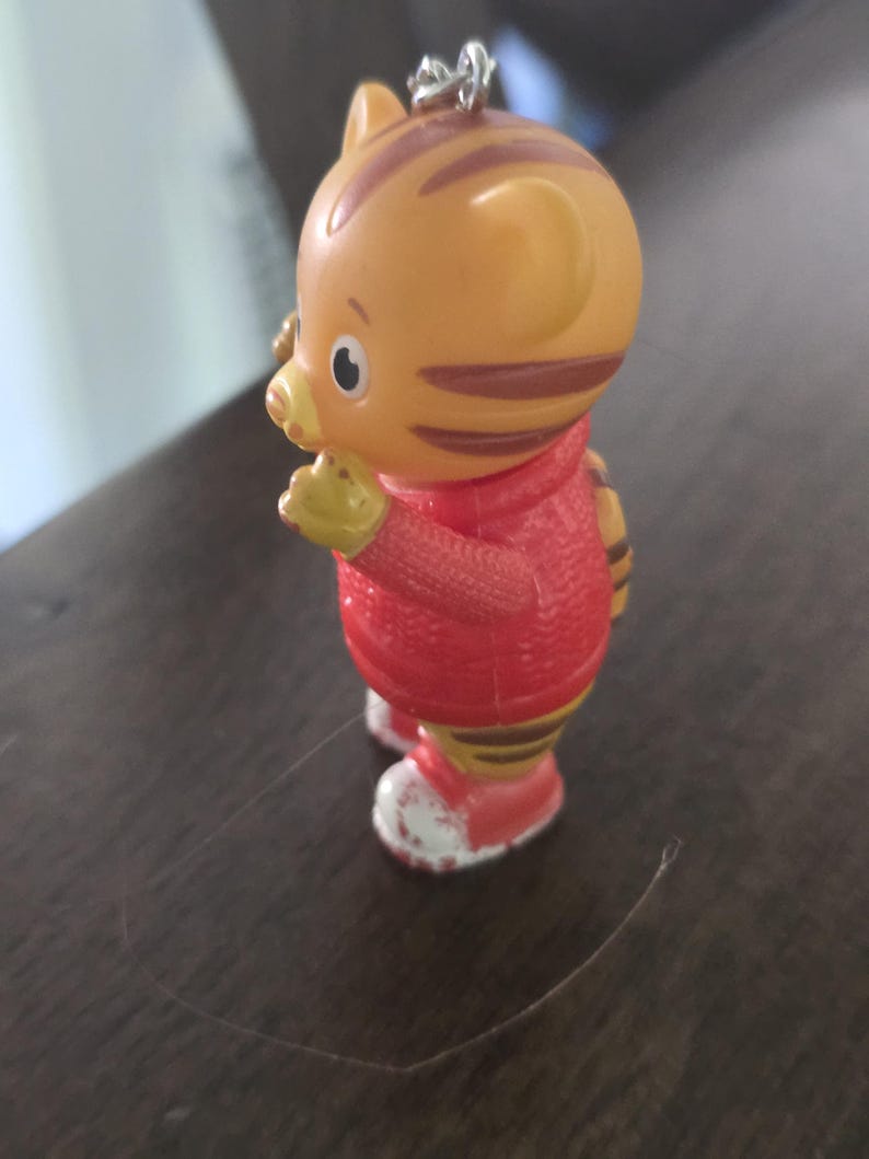 Op de afbeelding: Een klein oranje tijgerfiguurtje met bruine strepen, gekleed in een rode trui en rode schoenen met witte accenten. Het personage heeft een ronde kop, puntige oren en een metalen ring aan de bovenkant van het hoofd voor gebruik als sleutelhanger.