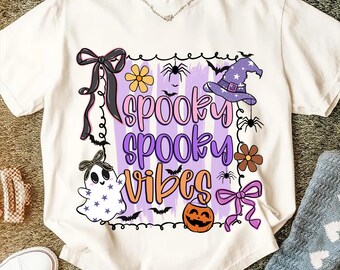 以下が含まれることがあります： ハロウィーンをテーマにしたグラフィックが施されたクリーム色のTシャツ。「spooky spooky vibes」という言葉が遊び心のあるフォントでデザインされており、お化け、魔女の帽子、クモ、コウモリ、花、カボチャ、リボンなどのイラストが描かれています。