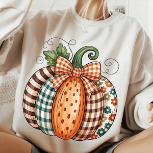 Peut inclure: Sweat-shirt blanc avec un motif de citrouille colorée. La citrouille est décorée de motifs à carreaux, à pois et floraux, ainsi que d'un nœud à carreaux. Le motif comprend des feuilles vertes et une vigne bouclée.