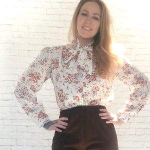 May include: A white blouse with a floral pattern in red, orange, and brown. The blouse features long sleeves, a bow tie collar, and is paired with dark brown pants. The outfit is set against a white brick wall.