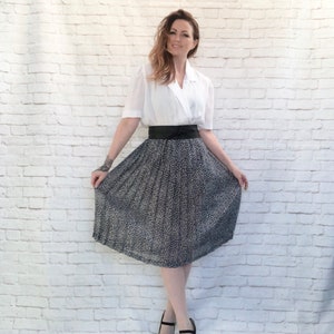 May include: A woman wearing a white blouse with a wrap-style front, a black belt, and a patterned skirt. The skirt has a dark background with a light-colored floral design. She is also wearing black heels.