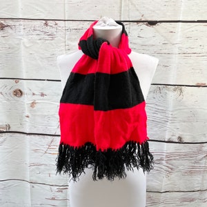 May include: A red and black striped scarf with black fringe. The scarf is displayed on a white mannequin. The scarf has alternating wide stripes of red and black. The fringe is at the bottom of the scarf.