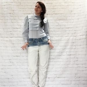 May include: A light blue blouse with layered detailing and long sleeves paired with two-tone jeans. The jeans transition from a dark blue waist to a white lower half. The model is wearing brown open-toe heels.