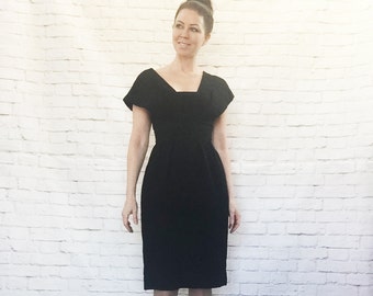 May include: A black dress with a square neckline and cap sleeves. The dress is knee-length and fitted, with a defined waist. The person is wearing black peep-toe heels. The background is a white brick wall.