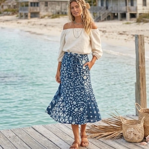Op de afbeelding: Een vrouw draagt een crèmekleurige off-the-shoulder top en een blauwe rok met bloemenpatroon. Ze staat op een houten pier bij de zee, met sandalen. De rok is ongeveer 76 cm lang.