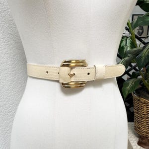 May include: Cream-colored belt with a gold-tone buckle. The belt has a textured, reptile-like pattern. The buckle is a large, rectangular shape with layered gold metal. The belt is displayed on a mannequin.