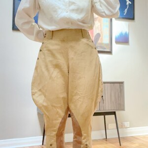 May include: Cream-colored button-down shirt paired with tan riding breeches. The breeches feature a high waist, rounded hips, and button details along the lower legs. The outfit suggests a vintage or equestrian theme.