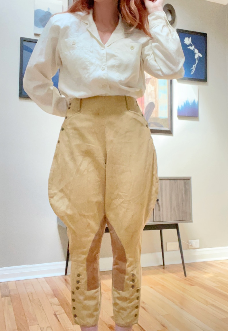 May include: Cream-colored button-down shirt paired with tan riding breeches. The breeches feature a high waist, rounded hips, and button details along the lower legs. The outfit suggests a vintage or equestrian theme.