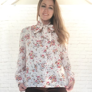 May include: A white blouse with a floral pattern in red, brown, and yellow. The blouse features long sleeves and a large bow at the neck. The background is a white brick wall.