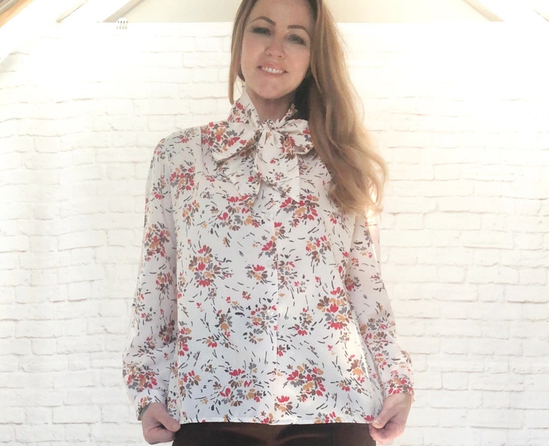 May include: A white blouse with a floral pattern in red, brown, and yellow. The blouse features long sleeves and a large bow at the neck. The background is a white brick wall.