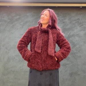 May include: A burgundy-colored, textured jacket with a matching scarf tied at the neck. The jacket has a soft, plush appearance and features pockets. The person is wearing a dark gray skirt. The background is a textured gray wall.