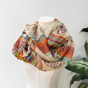 May include: An infinity scarf in a mix of patterns and stripes. The scarf features beige, orange, brown, and teal. The scarf is draped over a white mannequin, showcasing its design and texture.