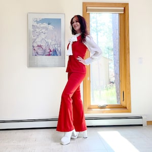 Puede incluir: Un traje pantalón rojo vintage con chaleco a juego y pantalones acampanados. El conjunto se combina con una blusa blanca y tacones de plataforma. Un póster enmarcado cuelga de la pared.