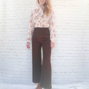 May include: A woman wearing a cream-colored blouse with a red floral pattern and a necktie. She is wearing dark brown, wide-leg pants and brown platform shoes. The outfit is set against a white brick wall.