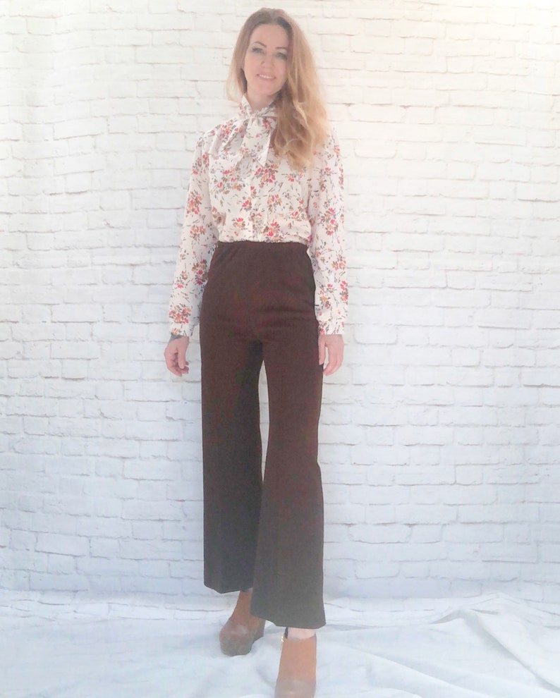 May include: A woman wearing a cream-colored blouse with a red floral pattern and a necktie. She is wearing dark brown, wide-leg pants and brown platform shoes. The outfit is set against a white brick wall.
