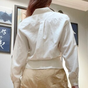 May include: A cream-colored button-down shirt with long sleeves. The shirt has a classic collar and a fitted waist. The sleeves are buttoned at the cuffs. The shirt is paired with a tan skirt. The background includes framed artwork.