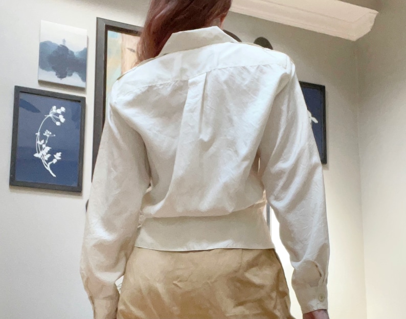 May include: A cream-colored button-down shirt with long sleeves. The shirt has a classic collar and a fitted waist. The sleeves are buttoned at the cuffs. The shirt is paired with a tan skirt. The background includes framed artwork.