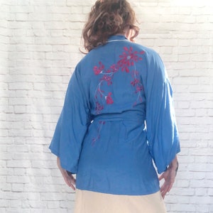 May include: A blue kimono-style robe with wide sleeves and a matching belt. The robe features a floral embroidery design in red on the back. The robe has a white trim around the collar.