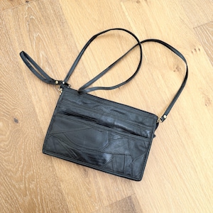 Op de afbeelding: Zwarte leren crossbodytas met een patchwork ontwerp aan de voorkant. De tas heeft een lange, verstelbare schouderriem en een polsbandje. De tas heeft meerdere vakken en een ritssluiting.