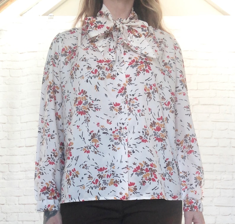 May include: A white blouse with a floral pattern in red, brown, and gray. The long-sleeved shirt features a large bow at the neckline. The blouse is a vintage style.