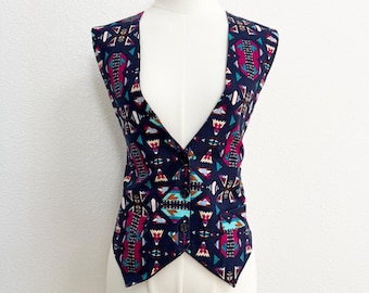 May include: A navy blue vest with a colorful geometric pattern. The vest features a V-neck, button closure, and two small pockets. The pattern includes teal, magenta, and white shapes.