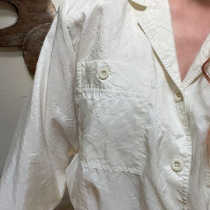 May include: Off-white button-down shirt with a collared neckline and a single chest pocket. The long-sleeved shirt has a textured, slightly wrinkled appearance. The buttons are round and match the shirt's color. The shirt is made of a lightweight material.