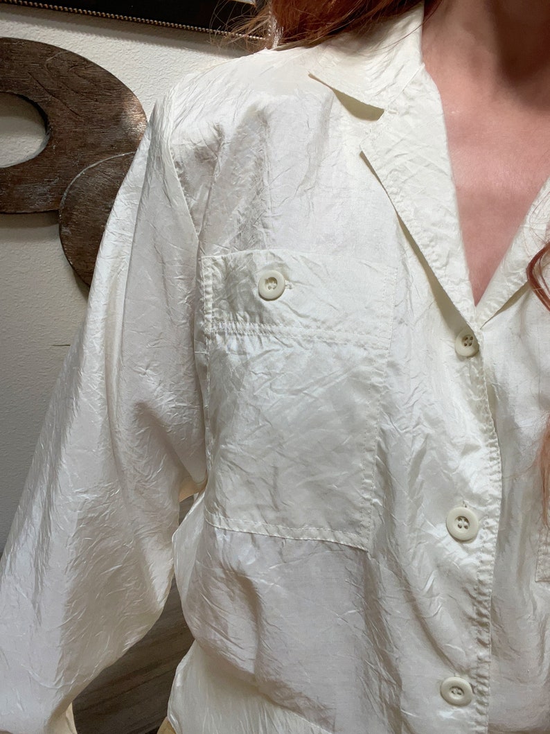 May include: Off-white button-down shirt with a collared neckline and a single chest pocket. The long-sleeved shirt has a textured, slightly wrinkled appearance. The buttons are round and match the shirt's color. The shirt is made of a lightweight material.