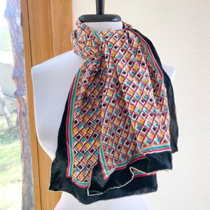 May include: A colorful scarf with a geometric pattern of squares in red, yellow, teal, and black on a cream background. The scarf has a black border and is draped over a white mannequin.