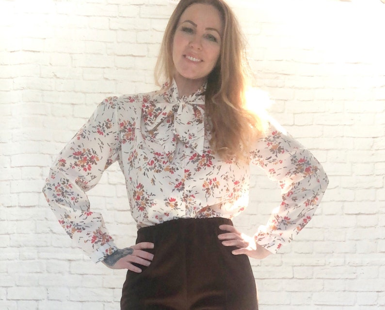 May include: A white blouse with a floral pattern in red, orange, and brown. The blouse features long sleeves, a bow tie collar, and is paired with dark brown pants. The outfit is set against a white brick wall.