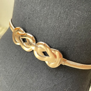 Puede incluir: Cinturón de metal dorado con una serie de eslabones entrelazados en forma de nudo. El cinturón tiene un diseño flexible en forma de resorte y se muestra sobre un fondo negro. El cinturón es un accesorio elegante.