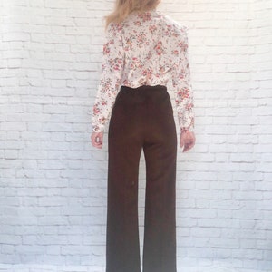 May include: A vintage outfit featuring a floral print blouse and wide-leg brown pants. The blouse has a white background with a colorful floral pattern. The pants are a solid, dark brown color, creating a retro aesthetic.