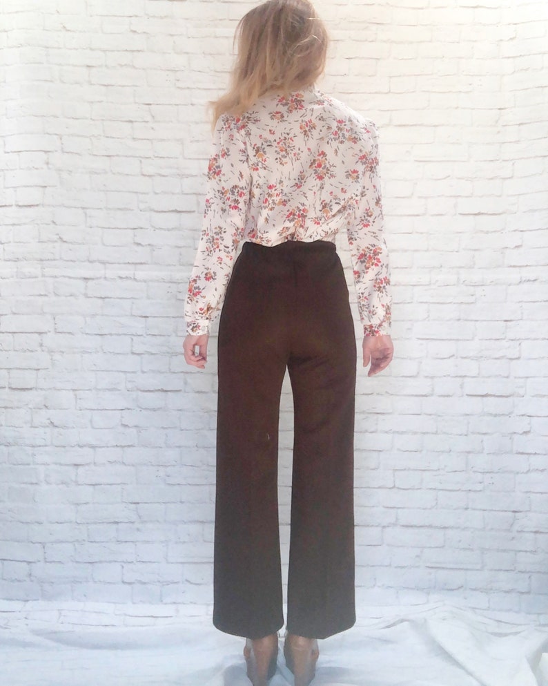 May include: A vintage outfit featuring a floral print blouse and wide-leg brown pants. The blouse has a white background with a colorful floral pattern. The pants are a solid, dark brown color, creating a retro aesthetic.