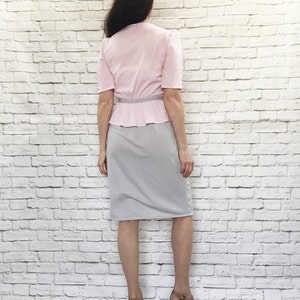 May include: A vintage two-piece outfit featuring a light pink blouse with short sleeves and a peplum waist, paired with a gray pencil skirt. The outfit is accessorized with a thin silver belt. The model is wearing platform heels.