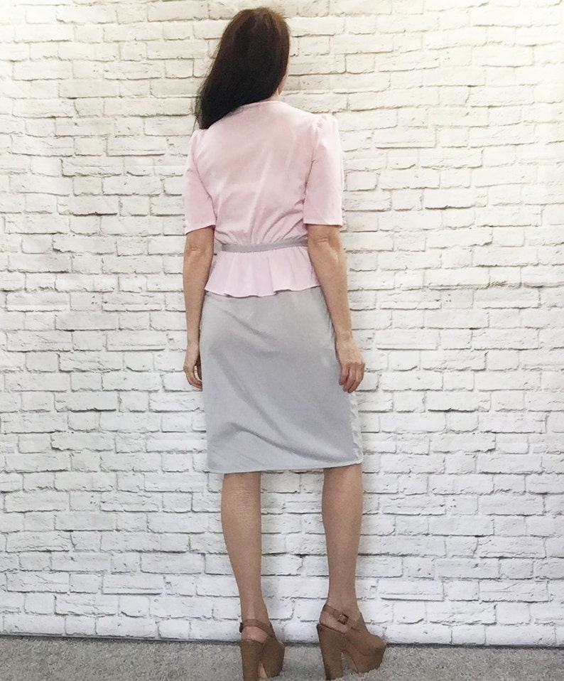 May include: A vintage two-piece outfit featuring a light pink blouse with short sleeves and a peplum waist, paired with a gray pencil skirt. The outfit is accessorized with a thin silver belt. The model is wearing platform heels.