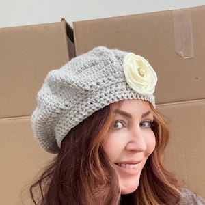May include: A light gray crocheted beret with a cream-colored flower accent. The flower has a pearl center. The beret is made of a soft, textured yarn. A stylish and elegant accessory for any outfit.