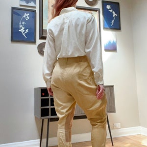 May include: A cream-colored long-sleeved button-down shirt paired with tan riding pants. The pants have a high waist, a fitted lower leg, and a suede-like patch on the outer thigh. The outfit is displayed indoors.